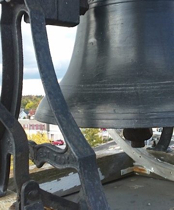 Colour photograph of the bell onto of steeple of current St. James Church. The bell is painted a grey colour. In the background is a view of the town of Fenelon Falls, with Cameron Lake in the very back of the picture.