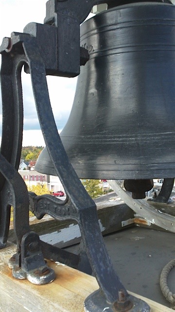 Colour photograph of the bell onto of steeple of current St. James Church. The bell is painted a grey colour. In the background is a view of the town of Fenelon Falls, with Cameron Lake in the very back of the picture.