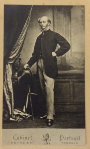 Circa 1850 photograph of a James Wallis, the first Minister and who provided land for the first St. James. Wallis was a tall man and is in this photograph is wearing a black waisted coat, with light coloured pants. He holds a cane and top hat in his right hand.