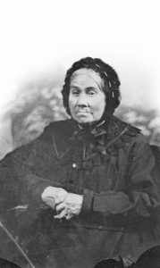 Sepia-tone photograph of an elderly woman, clad in a long black dress with a wide lace collar. Her white hair is tied in a bun behind her head and covered in a black lace bonnet (a béguin) tied with a large ribbon under her chin. Her hands, crossed on her lap, reveal fingers twisted and worn by old age.