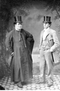 Image en noir et blanc de deux hommes se tenant debout dans un studio de photographie. L’un d’eux, un prêtre, est vêtu d’une soutane noire et s’appuie sur une canne à pommeau. L’autre homme porte un habit trois-pièces de couleur claire, ses mains sont dans les poches de son pantalon. Les deux hommes portent le chapeau haut de forme.