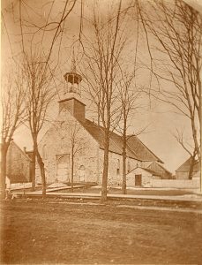 Photographie sépia de la première église de Saint-Jérôme. L’extérieur de l’église est en pierre des champs ainsi que le charnier situé à droite de l’église. Quelques petits bâtiments de ferme en bois se trouvent à droite de l’église. Un trottoir en bois longe une rue en terre devant le bâtiment.