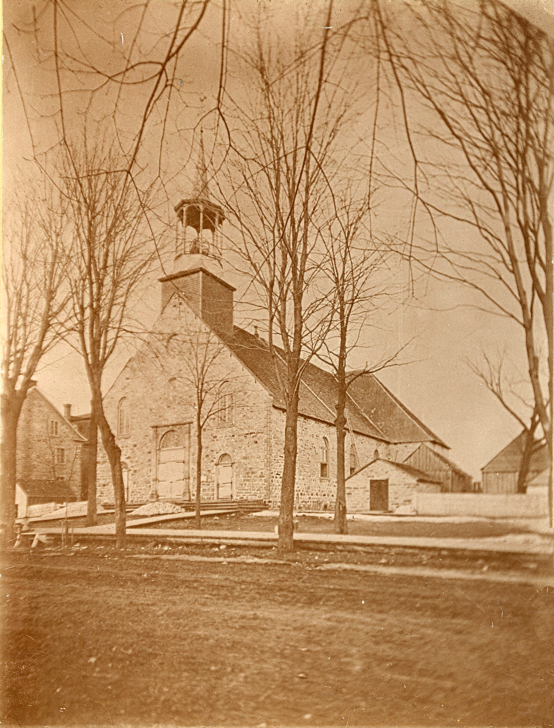 Sepia-tone photograph of the first Church in Saint-Jérôme. The church is built of fieldstone, as is the charnel house, visible to the right. Also seen at right are a number of small farm structures. A wooden sidewalk borders the dirt road in the foreground.