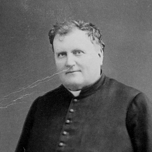 Photographie sépia d’un prêtre dans la quarantaine. Le portrait montre la partie supérieure de son corps. L’homme bien en chair, il a un double menton, porte une soutane et un col romain.