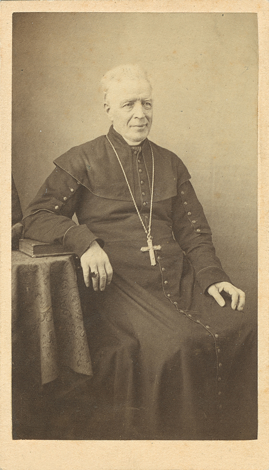 Sepia-tone photograph of a clergyman seated in a chair. He is elderly, with white hair. He wears a cassock, and a cross hangs from a long chain around his neck. His right hand rests on a table, with a ring visible on one finger.