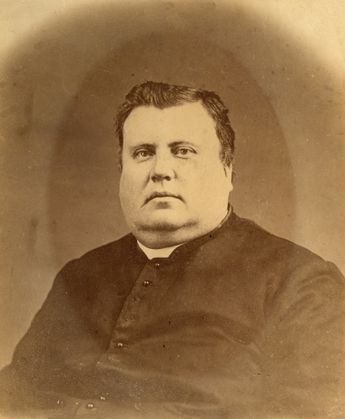 Photographie sépia d’un prêtre dans la quarantaine. Le portrait montre la partie supérieure de son corps. L’homme bien en chair, il a un double menton, porte une soutane et un col romain.