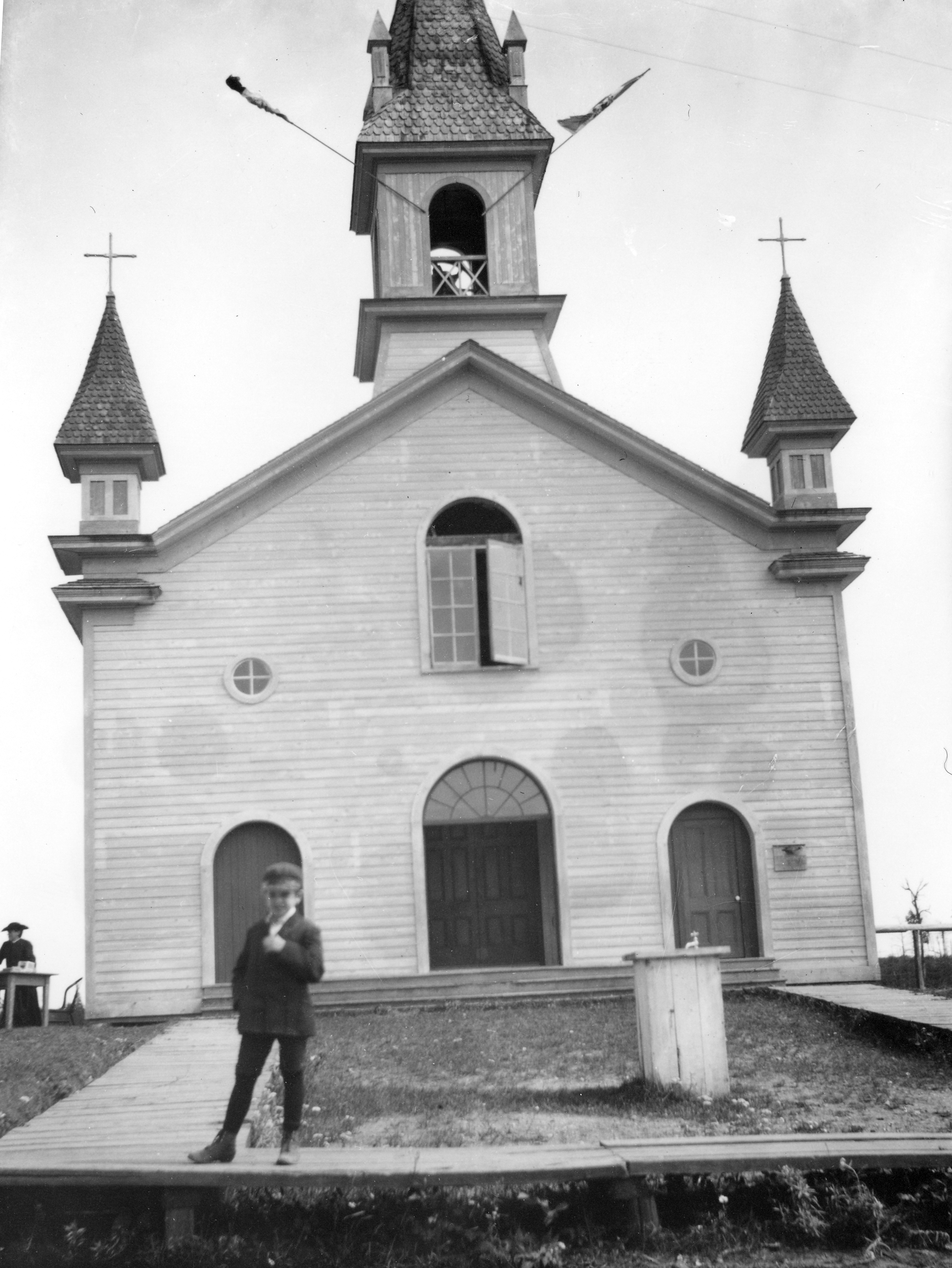 Black & white photograph of the façade of a small church. The clapboard building has three doors, three windows above the doors and a belfry. A boy stands on a boardwalk outside the church.