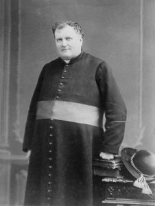 Black & white photograph of a man in his late fifties. He is standing and wears a cassock with a sash, symbolizing his status as Monseigneur. Two books and a felt hat rest on a low, carved table next to him. He rests his fist on the books.