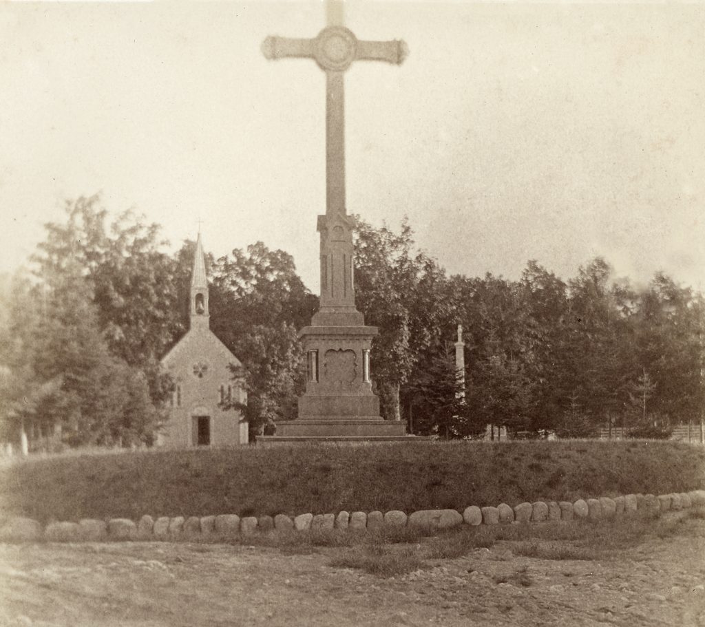 Photographie noir et blanc illustrant un cimetière en été. En avant-plan, une allée de sable bordant une plate-bande ornée d’une grande croix sculptée. En arrière-plan, une petite chapelle de bois coiffée d’un clocher entourée par quelques pierres tombales. Derrière la chapelle, la forêt.