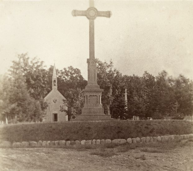 Black & white photograph depicting a cemetery in summertime. In the foreground is a sandy path, and behind it, a plant bed topped by a tall sculpted cross. Farther back, a small wooden chapel with a steeple is visible, surrounded by a few gravestones. In the background is a woodland.