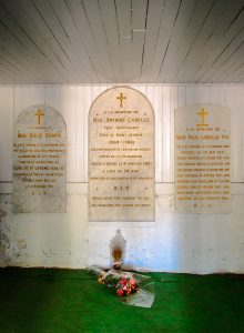 Colour photograph of a tomb in the basement of a chapel. Three white marble plaques with gold inscriptions are affixed to the wall. Beneath the central plaque, a bouquet of flowers and a glass urn site on the floor, which is carpeted in artificial grass.