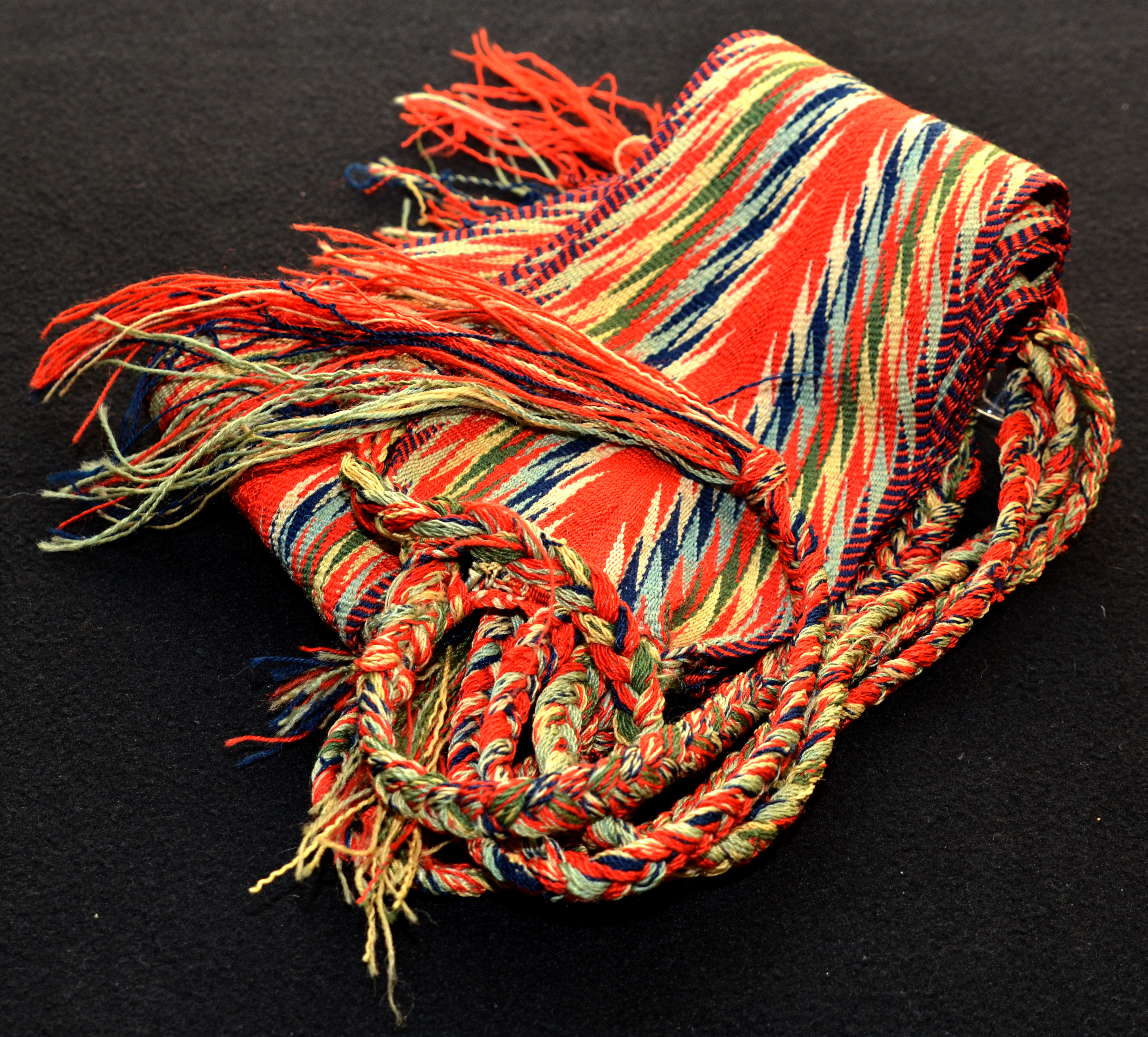 Colour photograph of an arrowhead sash, or ceinture fléchée, around 20 cm wide and 2 m long. The woven pattern looks like lightning bolts or arrowheads. The broad stripe down the middle is bright red, with symmetrical zigzags to the right and left of centre. On each end is a long fringe of three woollen braids.