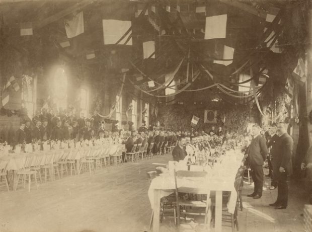 Sepia-tone photograph of a long, high-ceilinged reception hall, festooned with streamers, French flags and fir branches. Two long parallel tables are set for a meal, with several men seated at them. Members of a brass band stand against the wall to the left.