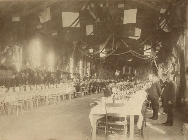 Sepia-tone photograph of a long, high-ceilinged reception hall, festooned with streamers, French flags and fir branches. Two long parallel tables are set for a meal, with several men seated at them. Members of a brass band stand against the wall to the left.