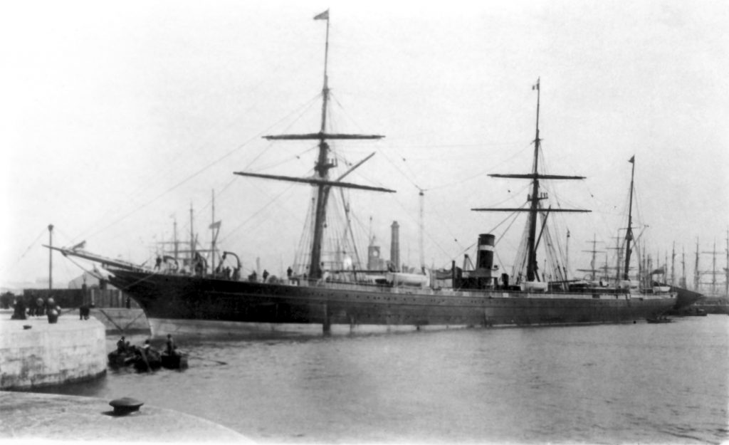 Black & white photograph of a sail and steam ship moored at a concrete dock in a port. The very large vessel sports three masts and a smokestack. A few rowboats are in the water nearby. In the background, masts of several other ships are seen.