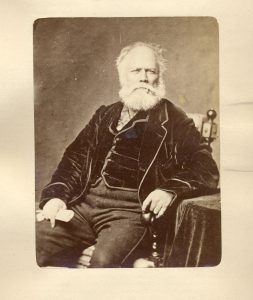 Photographie sépia d’un homme d’une soixantaine d’années assis sur une chaise. L’homme a les cheveux blancs, son front est dégarni. Il porte une barbe blanche fournie de longueur moyenne. Il est vêtu d’une veste et d’un gilet de velours foncé et d’un pantalon fabriqué dans un matériau plus léger. Un de ses bras est appuyé sur une table nappée de velours.