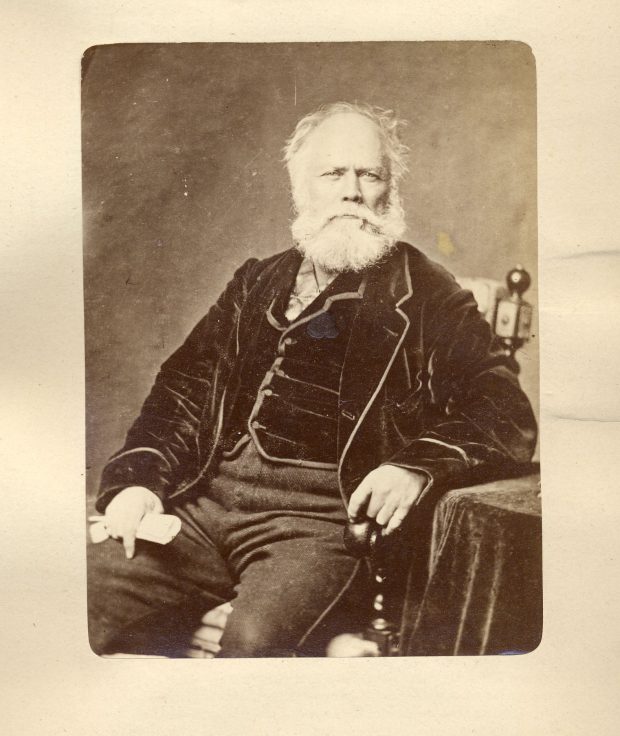 Sepia-tone photograph of a man aged about sixty, seated in a chair. His hair is white and he is slightly balding, with a thick, white, medium-length beard. He wears a dark velvet waistcoat and jacket, with trousers in a lighter material. His left arm rests on a table covered in a velvet tablecloth.