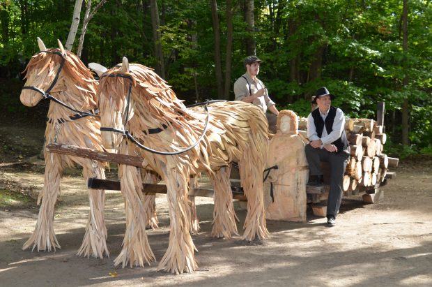 Colour photograph of a sculpture assembled from driftwood. The lifesize sculpture, located in a woodland, represents a sled filled with logs, pulled by two horses. A driver sculpted in wood sits at the front of the sled. Two men dressed in period costumes pose on the sled.