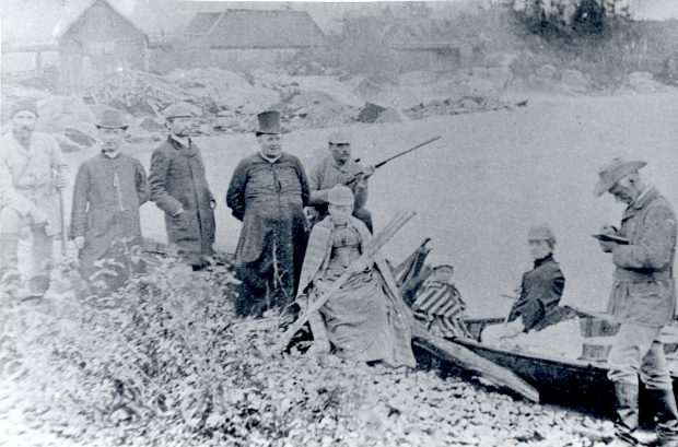 Black & white photograph of a group of people standing on a riverbank by a canoe: two priests, two women, a child, two men holding rifles, a man taking notes, and another man. Some wooden buildings and a wooden bridge are seen in the background.