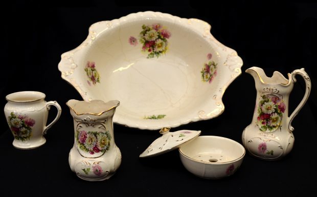 Colour photograph of a six-piece men’s toilet service in porcelain. In front, left to right: receptacle for items, shaving scuttle, soap dish, and water jug. At the rear: shaving bowl. The porcelain objects are white, with flower motifs and gilt edges.