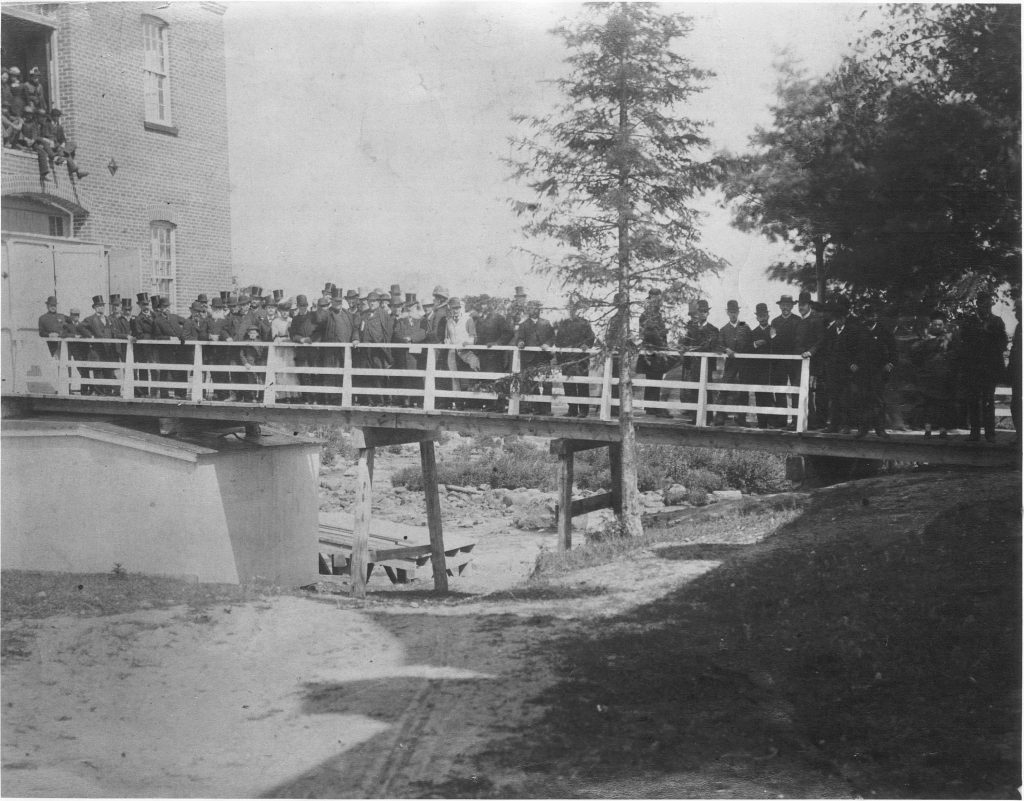 Black & white photograph of a group of people, mostly men, standing on a walkway in summer. The walkway leads to a large brick factory building. People are also seen perched higher up, in an opening on a higher floor of the factory. Many of those pictured are wearing top hats for the occasion.