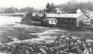 Photographie en noir et blanc d’un paysage au printemps. Au premier plan à droite, un petit pont de bois enjambant une rivière houleuse et un amoncellement billots de bois. En amont, un barrage d’où l’eau s’écoule avec force. Au centre, un bâtiment industriel de brique. En arrière-plan à gauche, une maison et quelques bâtiments de ferme.