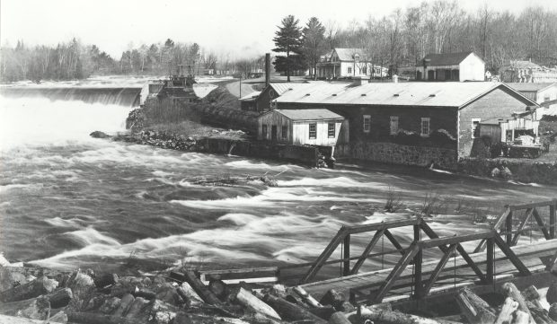 Black & white photograph of a landscape in spring. In the foreground at right, a small wooden bridge crosses a fast-flowing river with rapids; a pile of logs is also seen. Upstream, in the background at left, is a dam with water spilling over it forcefully. In the centre is an industrial brick building, and behind it, a house and a few farm buildings.