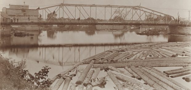 Sepia-tone photograph of an iron bridge and river in summer. People can be seen on the bridge deck and on the girders up above. A log boom is visible in the foreground, with some wooden buildings in the background.