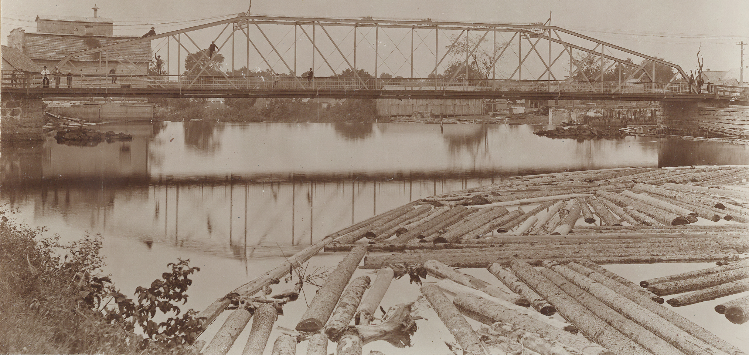 Sepia-tone photograph of an iron bridge and river in summer. People can be seen on the bridge deck and on the girders up above. A log boom is visible in the foreground, with some wooden buildings in the background.