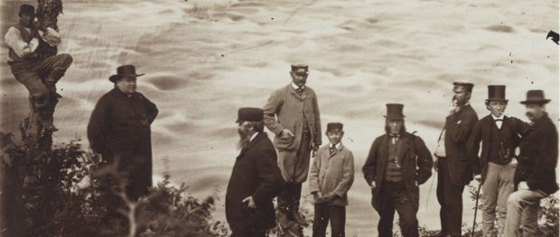 Black & white photograph of a group of nine people standing on the shore of a rushing river. At left, a man is perched partway up a tree trunk. Curé Labelle, a heavy-set man, is seen wearing a cassock and a wide-brimmed felt hat. At centre and right are six men and a teenaged boy wearing three-piece suits and top hats, caps or felt hats. Two of them are smoking pipes.