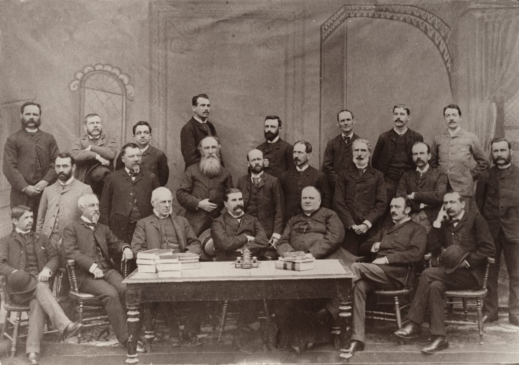 Black & white photograph depicting the 23 members of the Ministry of Agriculture and Colonization on September 1, 1889. Sixteen men stand behind group of a seven seated on chairs behind a table.