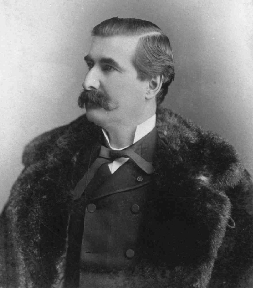 Black & white photograph of a man in his forties, with dark hair and a thick moustache. He wears elegant clothes beneath a fur coat.