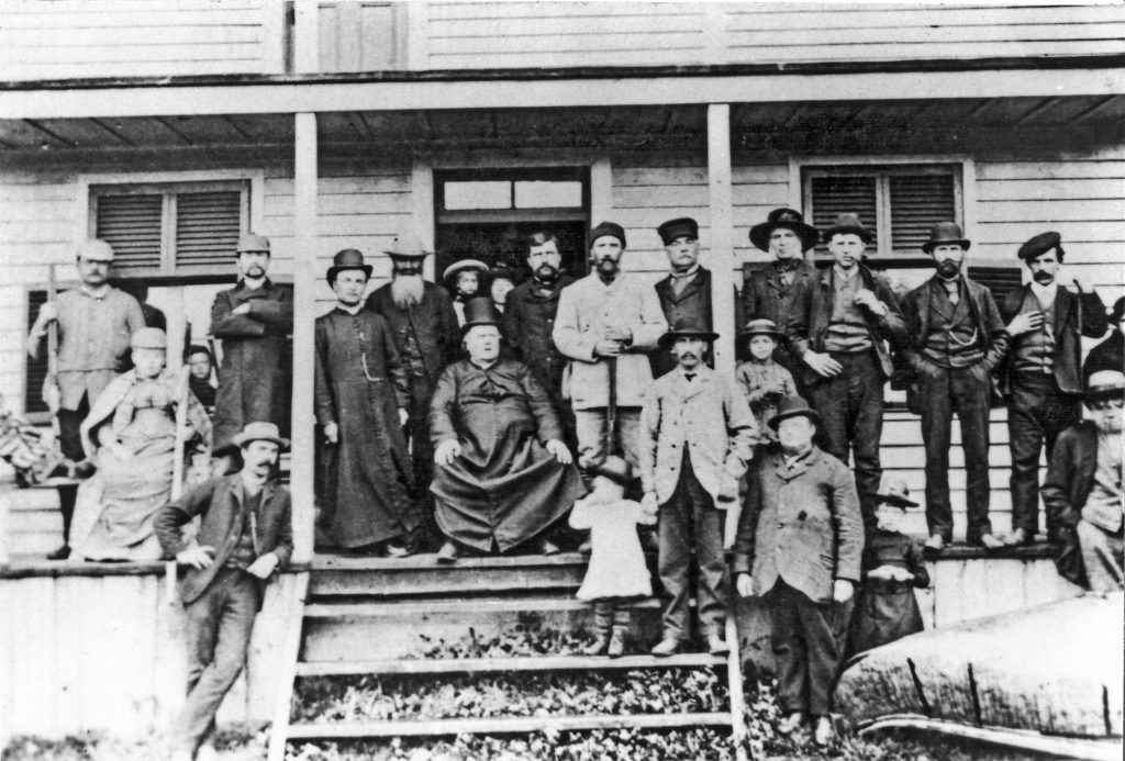 Black & white photograph of a group of around twenty people on the porch of a wooden house. In the centre of the group, Curé Labelle, a heavy-set man wearing a cassock, is seated in a chair. At right, on the ground in front of the porch, is a large birchbark canoe.