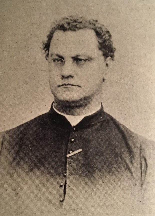 Sepia-tone photograph of a man in his thirties. He has close-cropped dark curly hair. He wears small oval eyeglasses. He wears a cassock, with a clerical collar.
