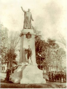 Photographie noir et blanc de la statue du curé Labelle qui pointe le doigt en direction du nord. La statue repose sur un imposant socle. Aux pieds du curé Labelle, on retrouve ses armoiries et une statue plus petite en bronze d’un paysan. Un homme et des enfants se trouvent près du socle. 