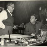 Black & white photograph of a man standing beside a desk, talking to other man seated behind it. The desk is covered with many items: a large, open book, a stapler, a pair of glasses, an inkwell and pen, and many other small objects.