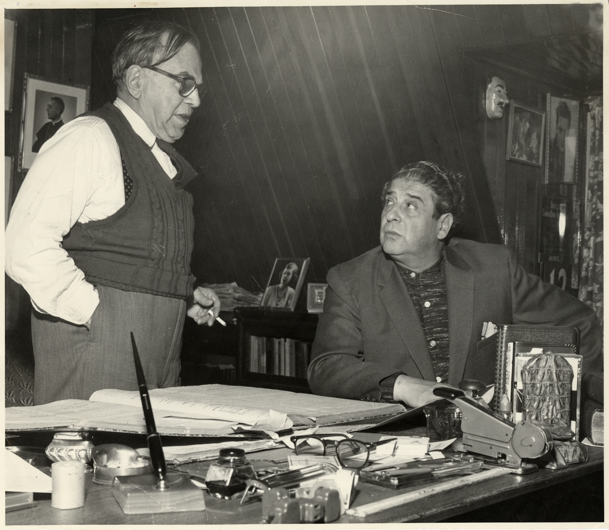 Black & white photograph of a man standing beside a desk, talking to other man seated behind it. The desk is covered with many items: a large, open book, a stapler, a pair of glasses, an inkwell and pen, and many other small objects.