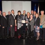 Photographie couleur de 14 hommes et femmes entourant la statue d’un ours, personnifiant le curé Labelle, déposée sur une table.