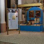 Photographie couleur de panneaux de textes et d’images d’une exposition entourant des vitrines d’objets.