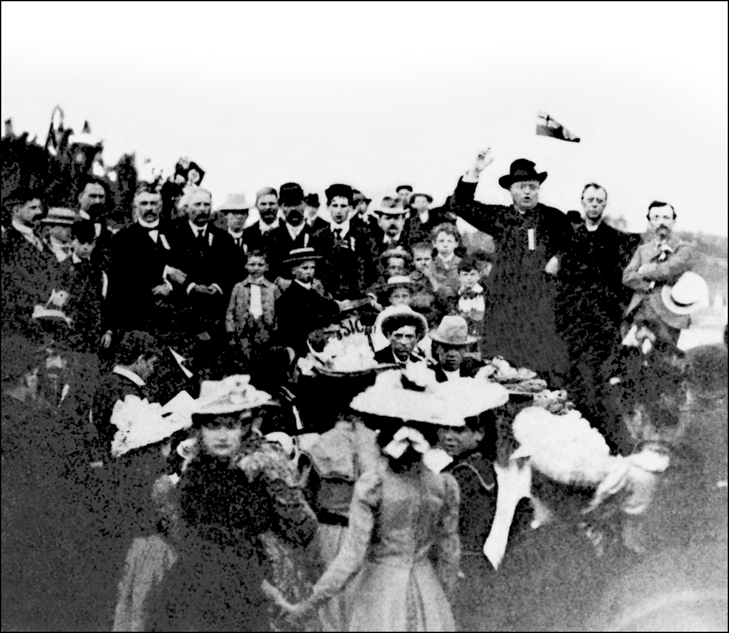 Black & white photograph of a crowd outdoors, surrounding an imposing figure, Curé Antoine Labelle. Several men are seen, along with a few women and children, all well dressed. Labelle wears a black cassock and black hat and is giving a speech; his mouth is open and his right arm is raised beside his head. His left art rests on his hip.