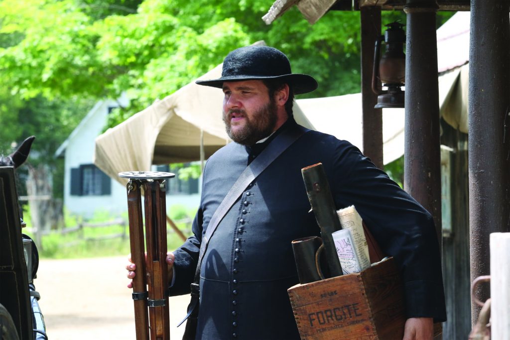 Colour photograph of an actor playing the role of Curé Labelle. He wears a cassock and a hat, and carries a box of maps.