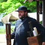 Colour photograph of an actor playing the role of Curé Labelle. He wears a cassock and a hat, and carries a box of maps.