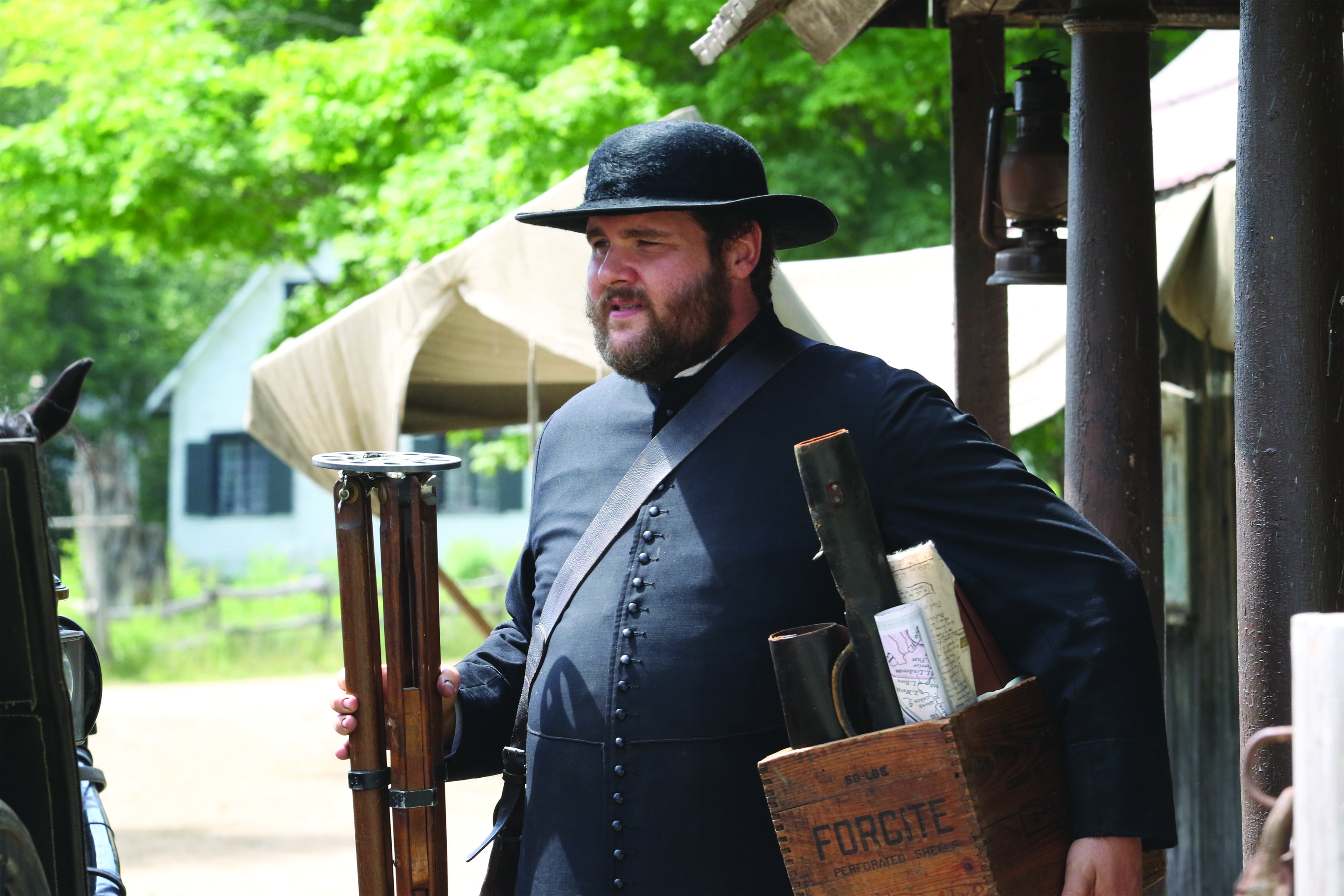 Colour photograph of an actor playing the role of Curé Labelle. He wears a cassock and a hat, and carries a box of maps.