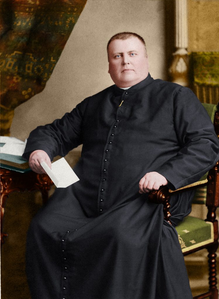 Colourized photograph of Curé Labelle seated at a desk, holding a letter. His right hand rests on the desk, and his left hand rests on the arm of his chair.