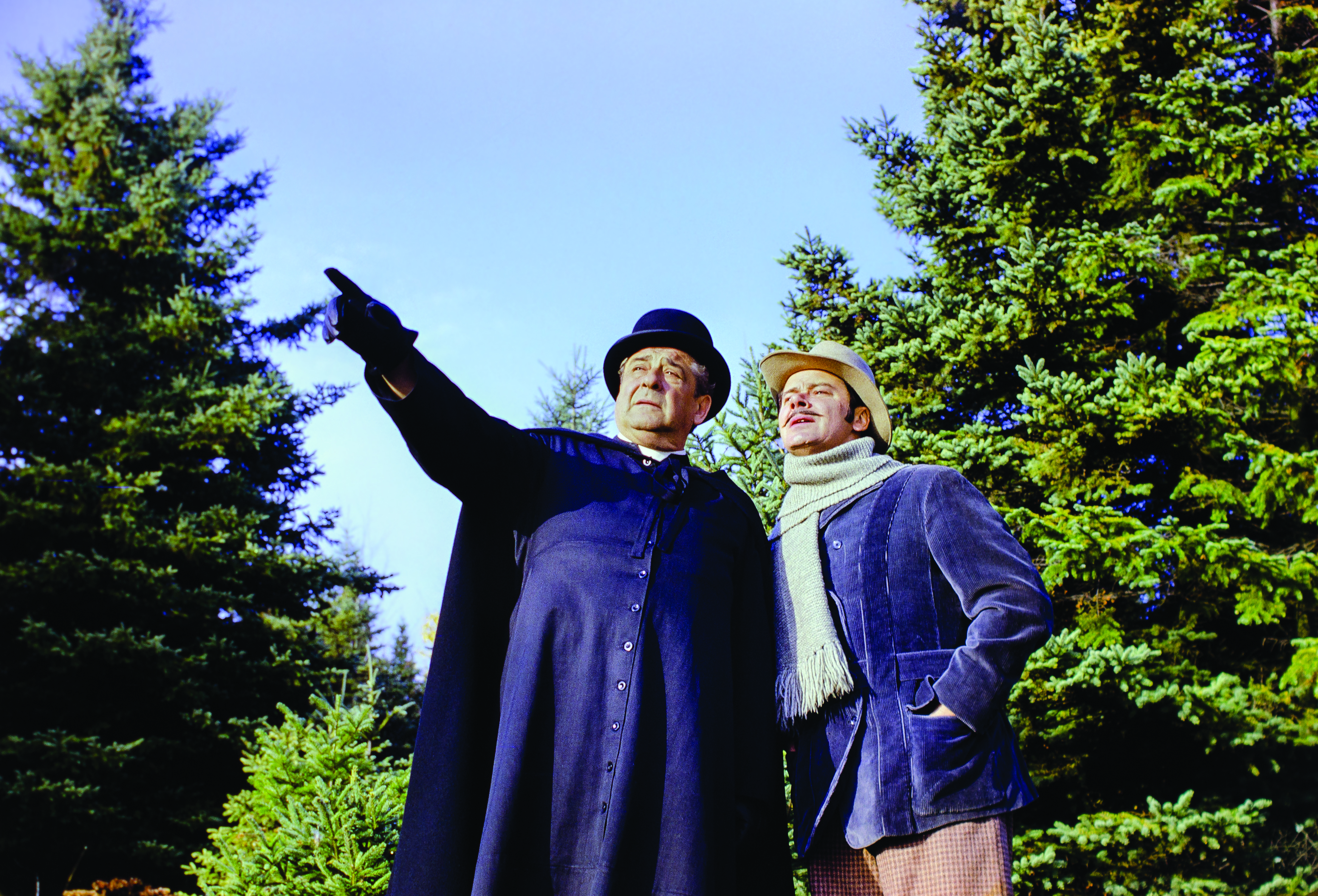 Colour photograph of two actors in a forest setting. A man in a cassock, playing Curé Labelle, points to something in the distance. The man accompanying him, wearing a coat, scarf and hat, stands close to him and gazes into the distance.