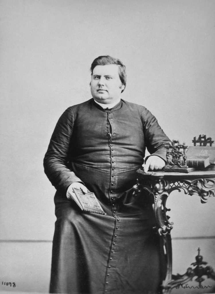 Black & white photographic portrait of a man aged thirty, fairly stout and with dark hair. He is seated, clad in a black cassock buttoned to the neck; his left arm rests on an ornately carved table and in his right hand he holds a book. His gaze is neutral and he is unsmiling.