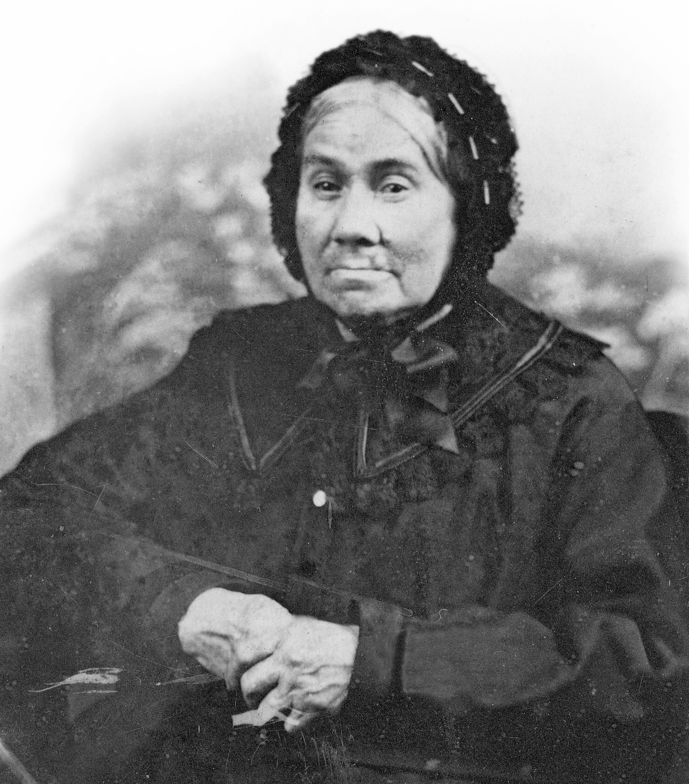 Sepia-tone photograph of an elderly woman, clad in a long black dress with a wide lace collar. Her white hair is tied in a bun behind her head and covered in a black lace bonnet (a béguin) tied with a large ribbon under her chin. Her hands, crossed on her lap, reveal fingers twisted and worn by old age.