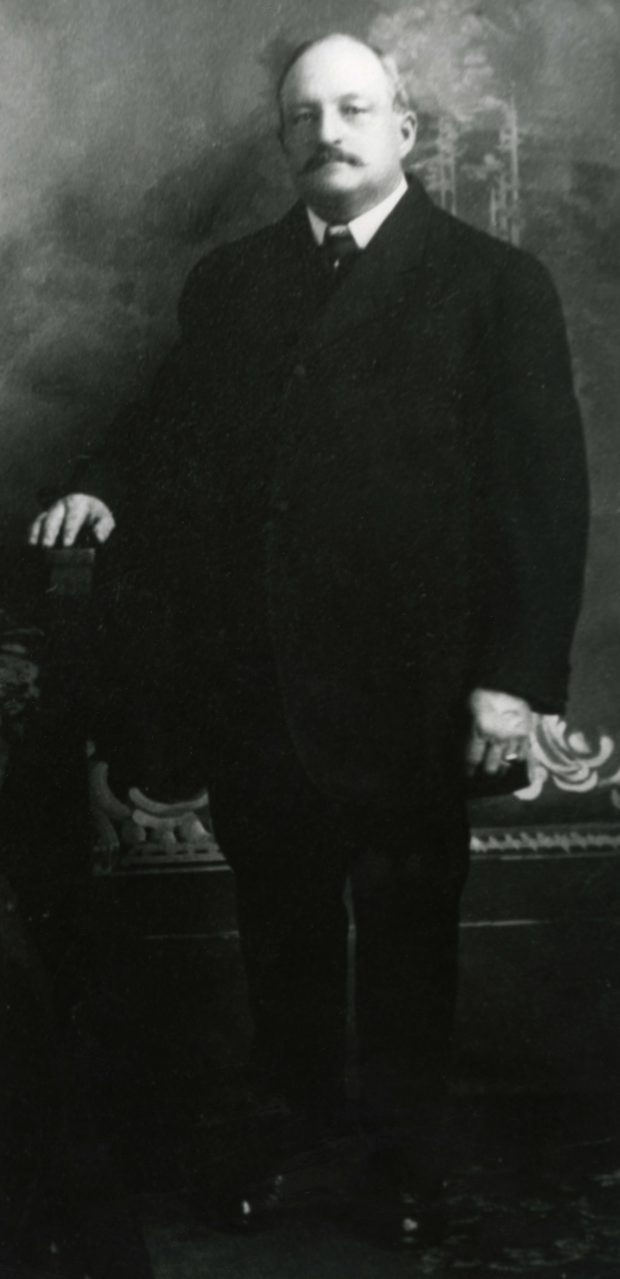 Old black and white photo of a man standing, wearing a black vest