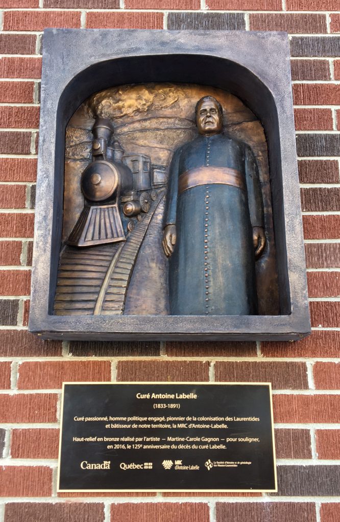 High-relief sculpture of Curé Labelle. On the left-hand side is a locomotive seen from the front, with train tracks; and on the right is Labelle, standing.