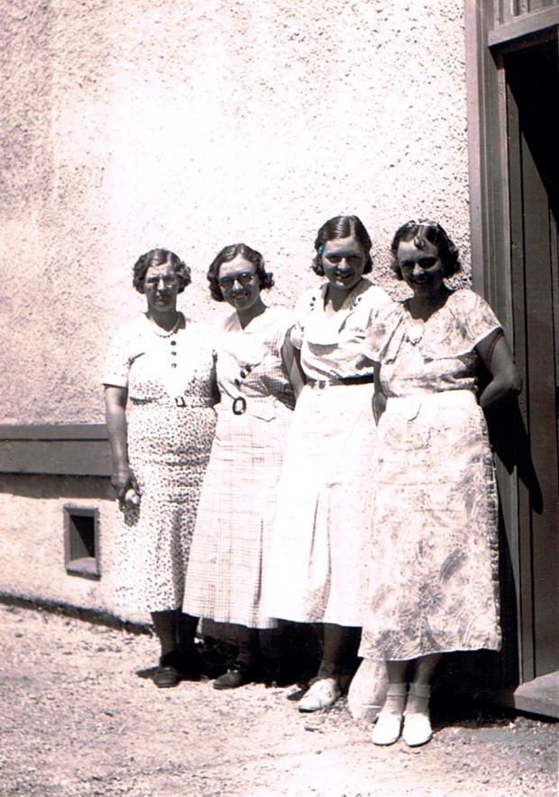 Black and white photo of four women standing outside a building. Each is wearing a dress and has a short, wavy hairstyle.